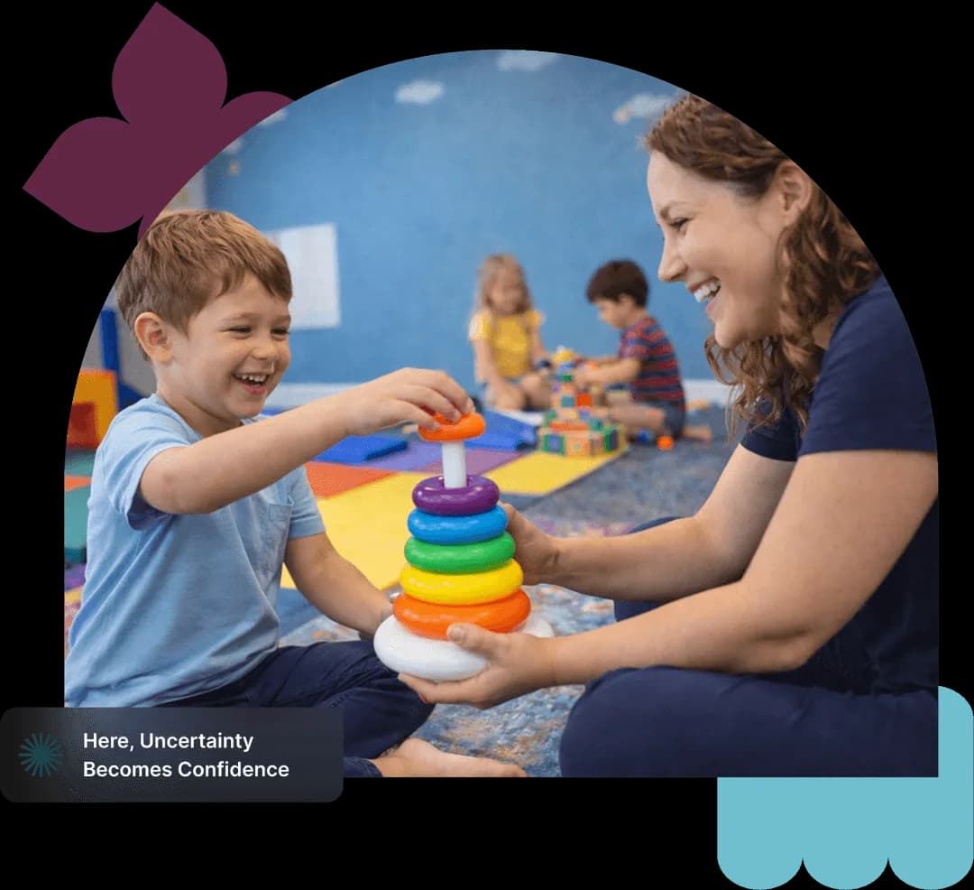 Smiling child and woman sitting on floor playing with a colorful stacking ring toy in a bright classroom.