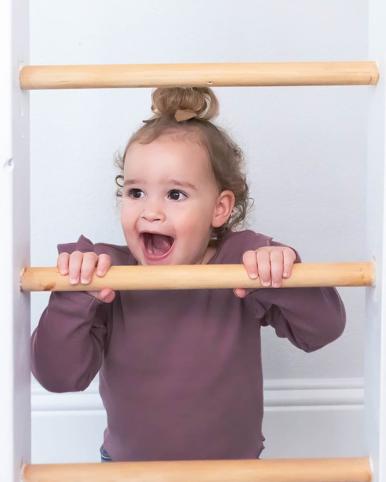 A young girl with curly hair in a bun holding onto wooden ladder rungs, smiling with her mouth open.