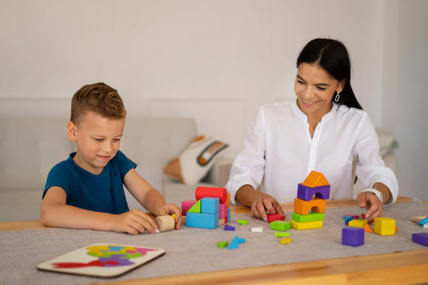 Woman and boy playing with colorful wooden blocks and a puzzle on a table indoors