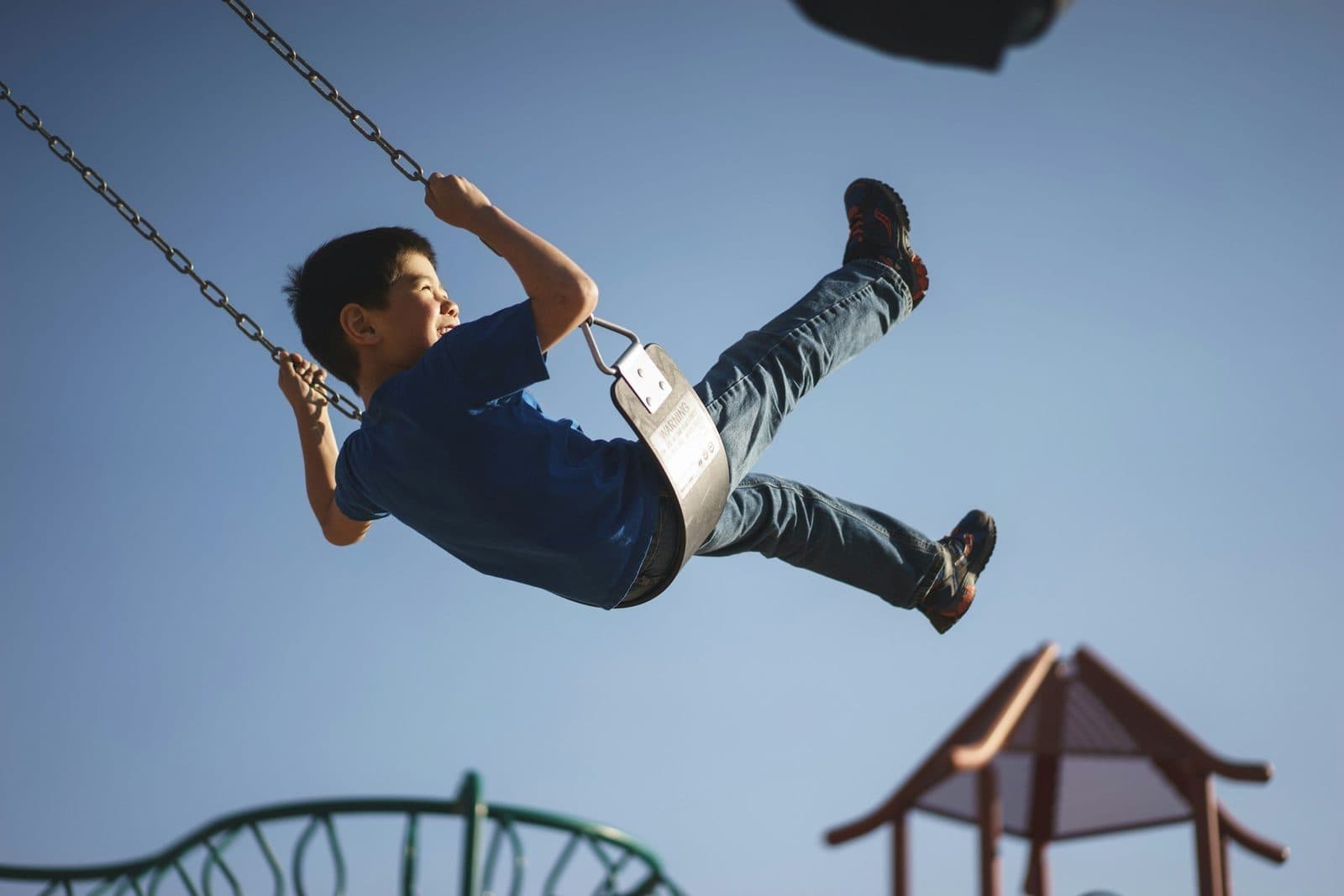 Child swinging high in the air at a playground against a clear blue sky.