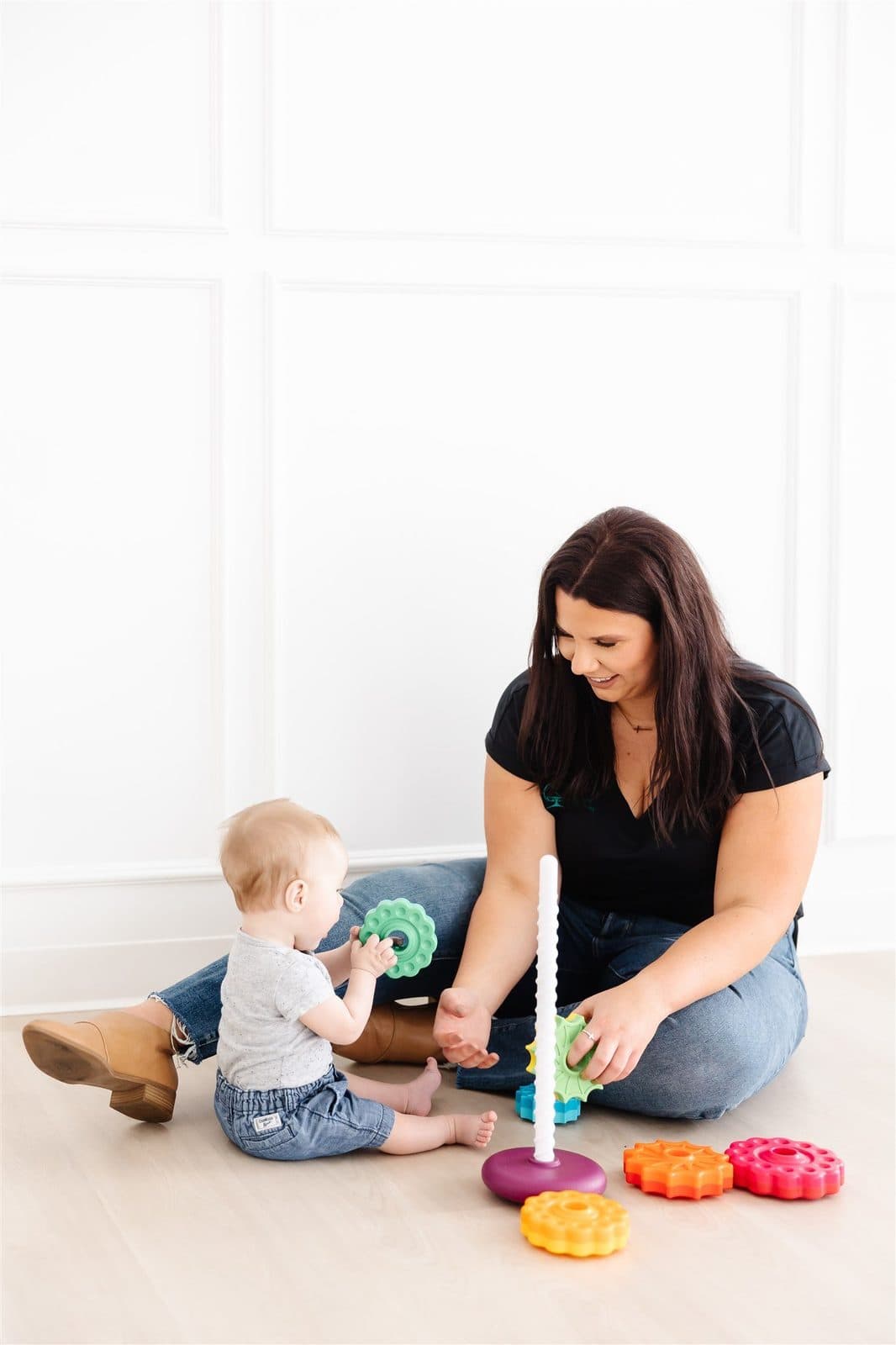 A woman and a baby playing with colorful stacking toys on the floor inside a bright room with white walls