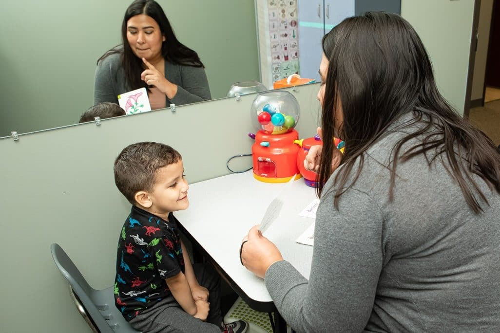Speech therapist working with a young boy at a table in a therapy room with a mirror, using picture cards and a gumball reward machine
