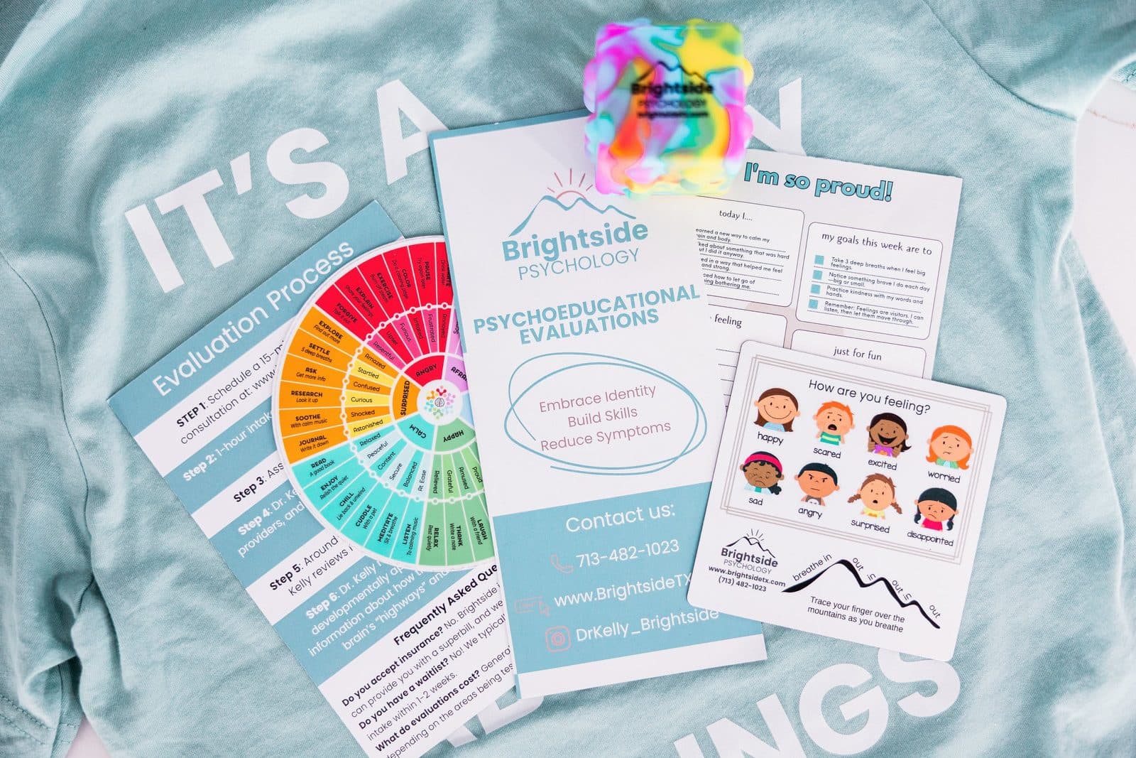 Various printed materials from Brightside Psychology, including evaluation forms, a rainbow-colored stress ball, and a card showing different emotions on a light blue fabric surface