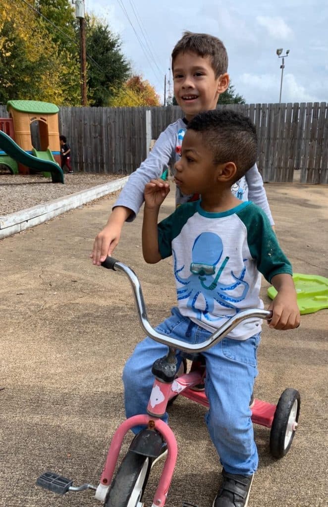 Two children playing together on a tricycle in the outdoor playground area at Building Greatness Autism Center