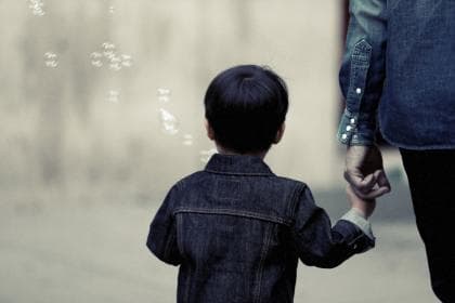 A young child in a denim jacket holding an adult's hand, looking at floating bubbles