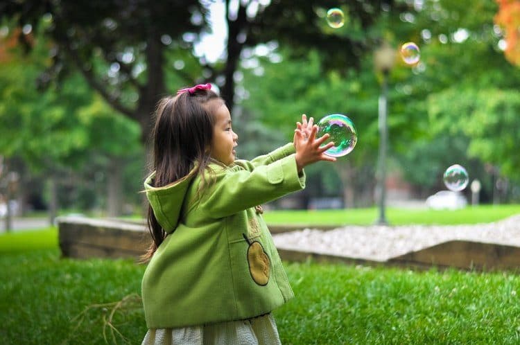 A young girl with long dark hair joyfully reaching out to catch floating bubbles in a grassy park