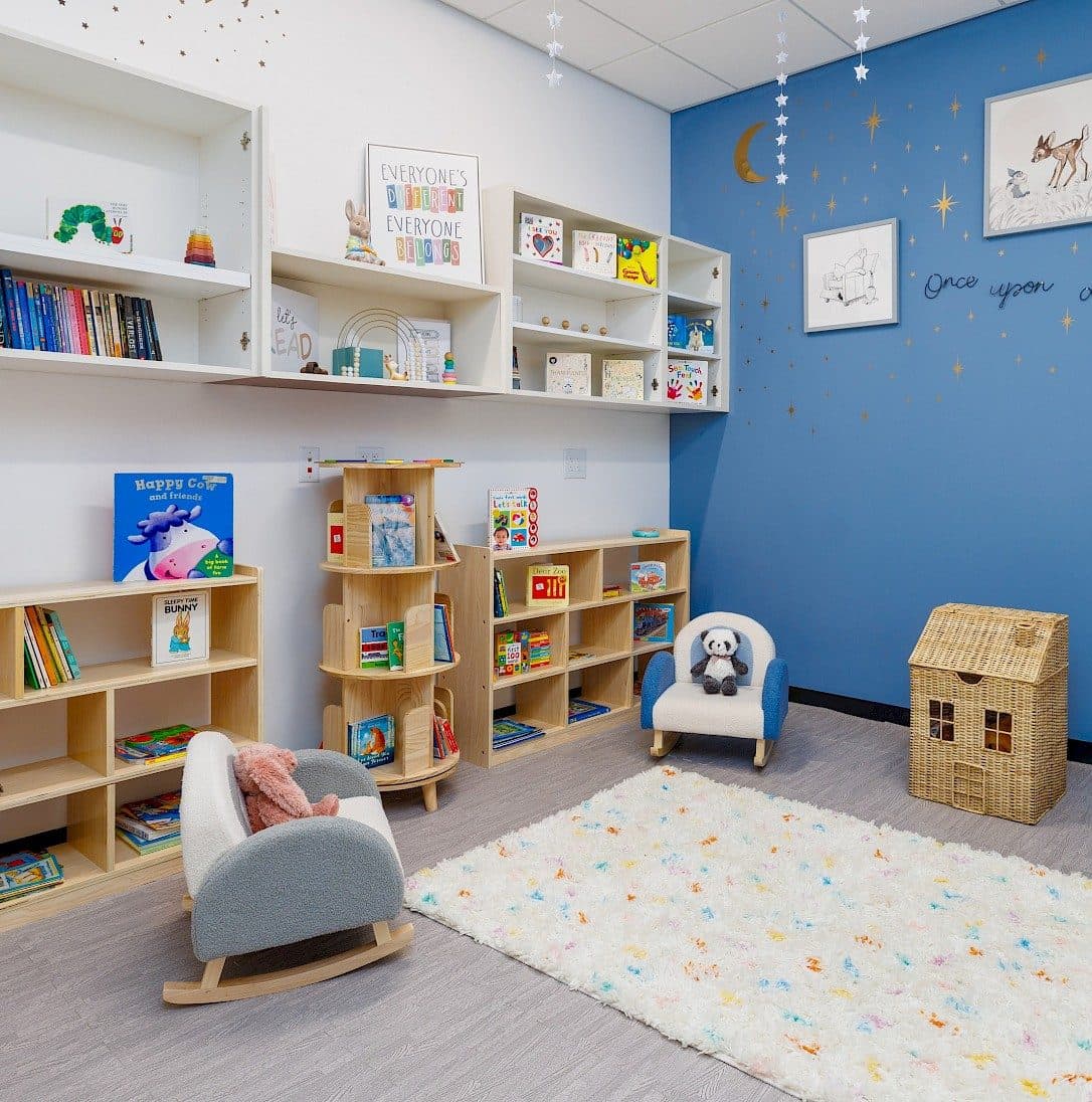 A cozy children's reading corner featuring bookshelves, rocking chairs, and a soft rug under a starry-themed wall.
