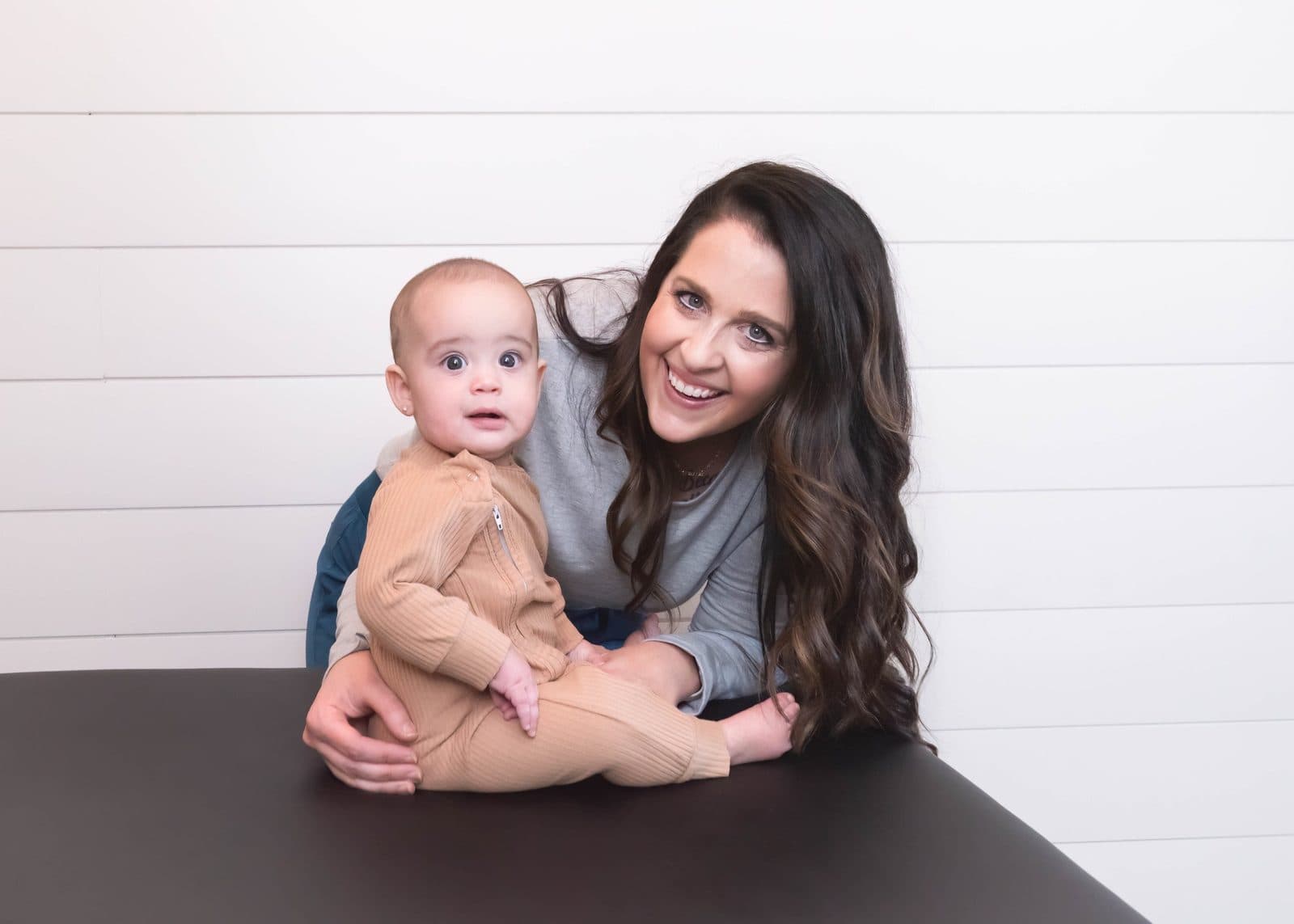 A woman with long wavy brown hair smiling while holding a young child with wide eyes, sitting at a black table against a white paneled wall.
