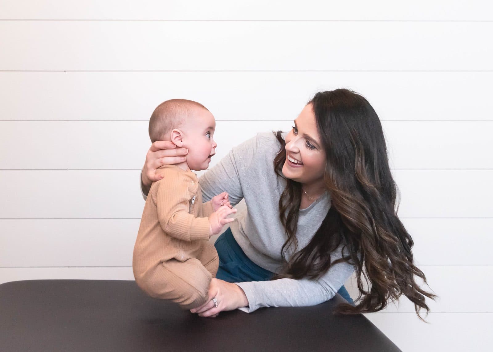 A woman with long dark hair and a gray shirt smiles while holding a baby who is sitting on a black table, with a white, shiplap wall behind them.