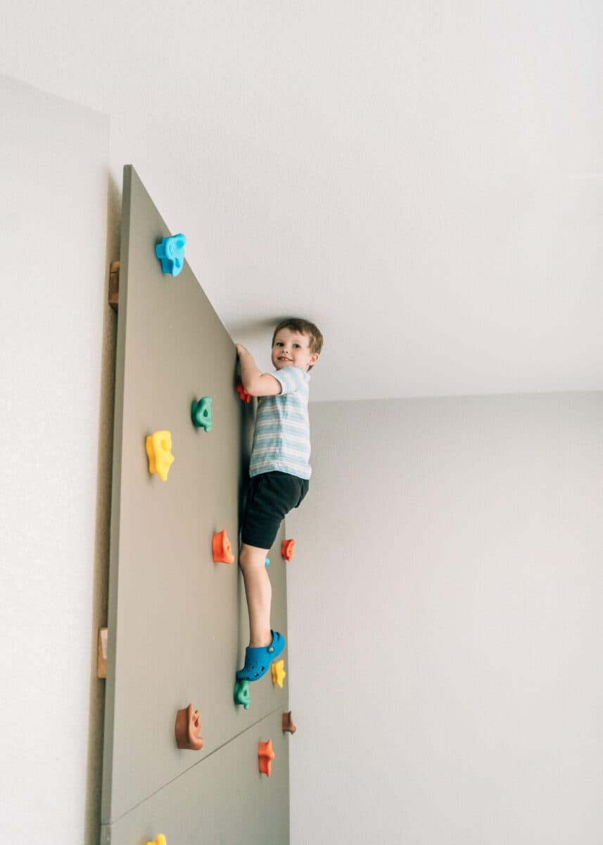 A young child climbs an indoor rock wall with colorful handholds during a gross motor activity.