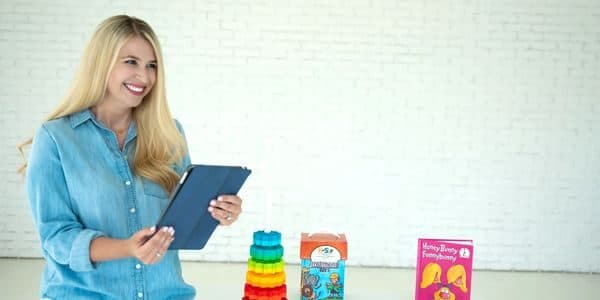 Smiling woman holding a tablet with colorful toys and books on a table.