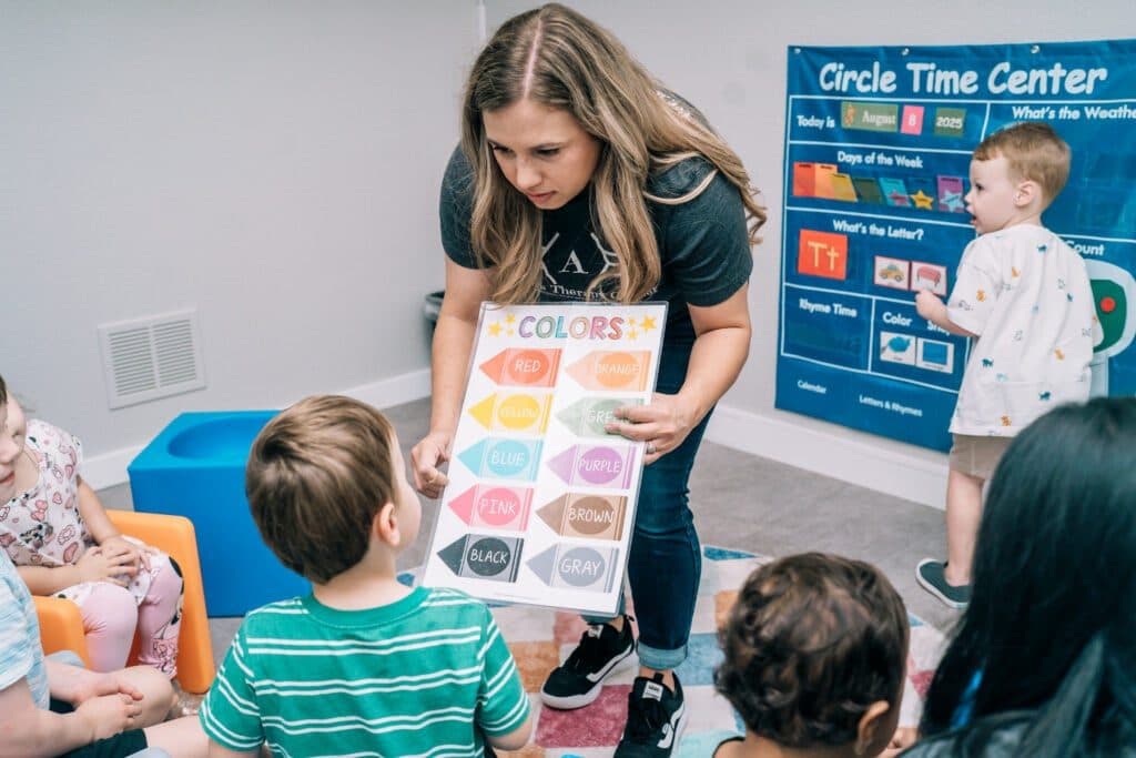 A therapist shows a colors chart to children seated on the floor during a group learning activity.