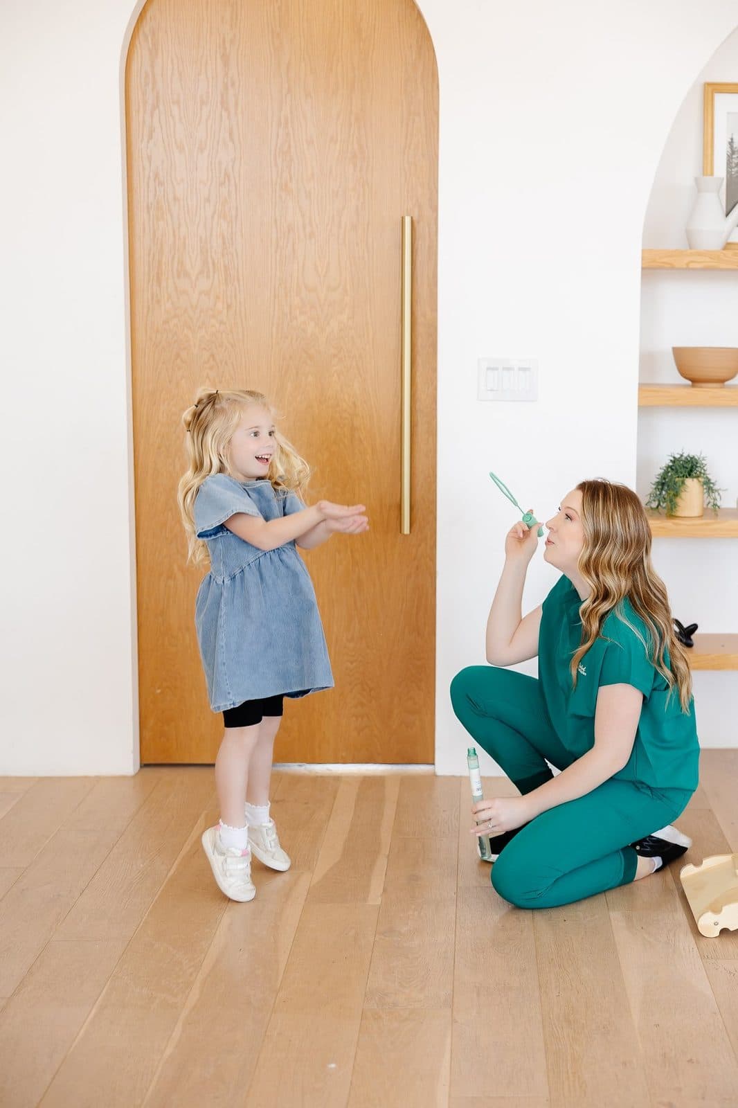 A young girl and a woman, likely a healthcare worker, are indoors. The girl is smiling and clapping her hands