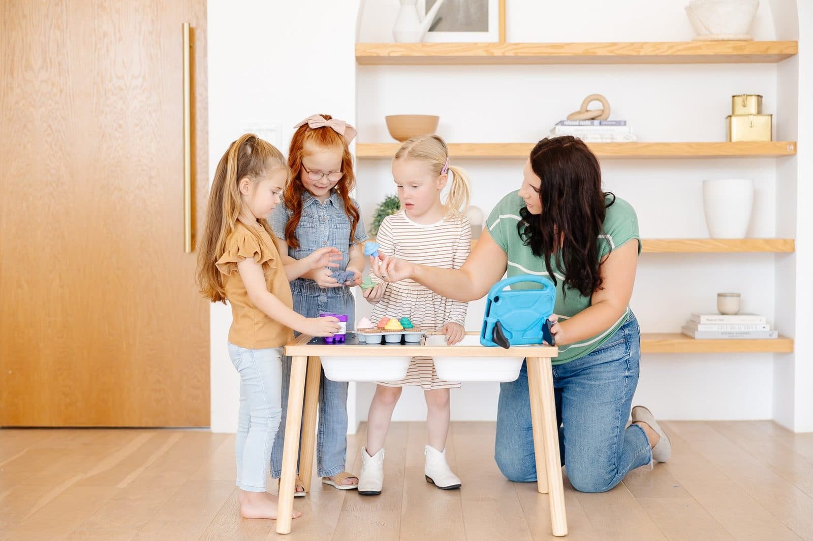 A woman kneeling at a small wooden table with four young girls playing with toy baking supplies