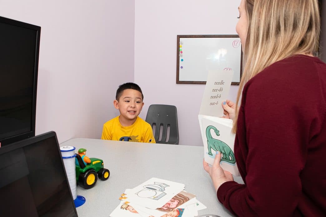 Therapist holding a phonics card book while a child responds across the table in a therapy room with whiteboard, toy tractor, and flashcards