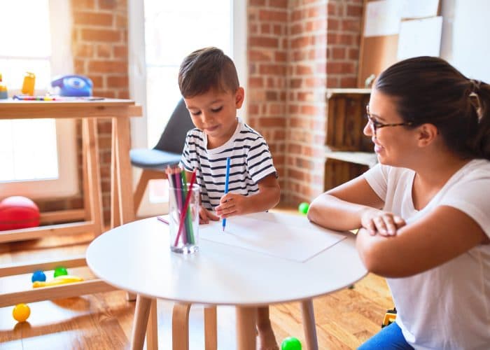 Beautiful teacher and toddler boy drawing with colored pencils at kindergarten